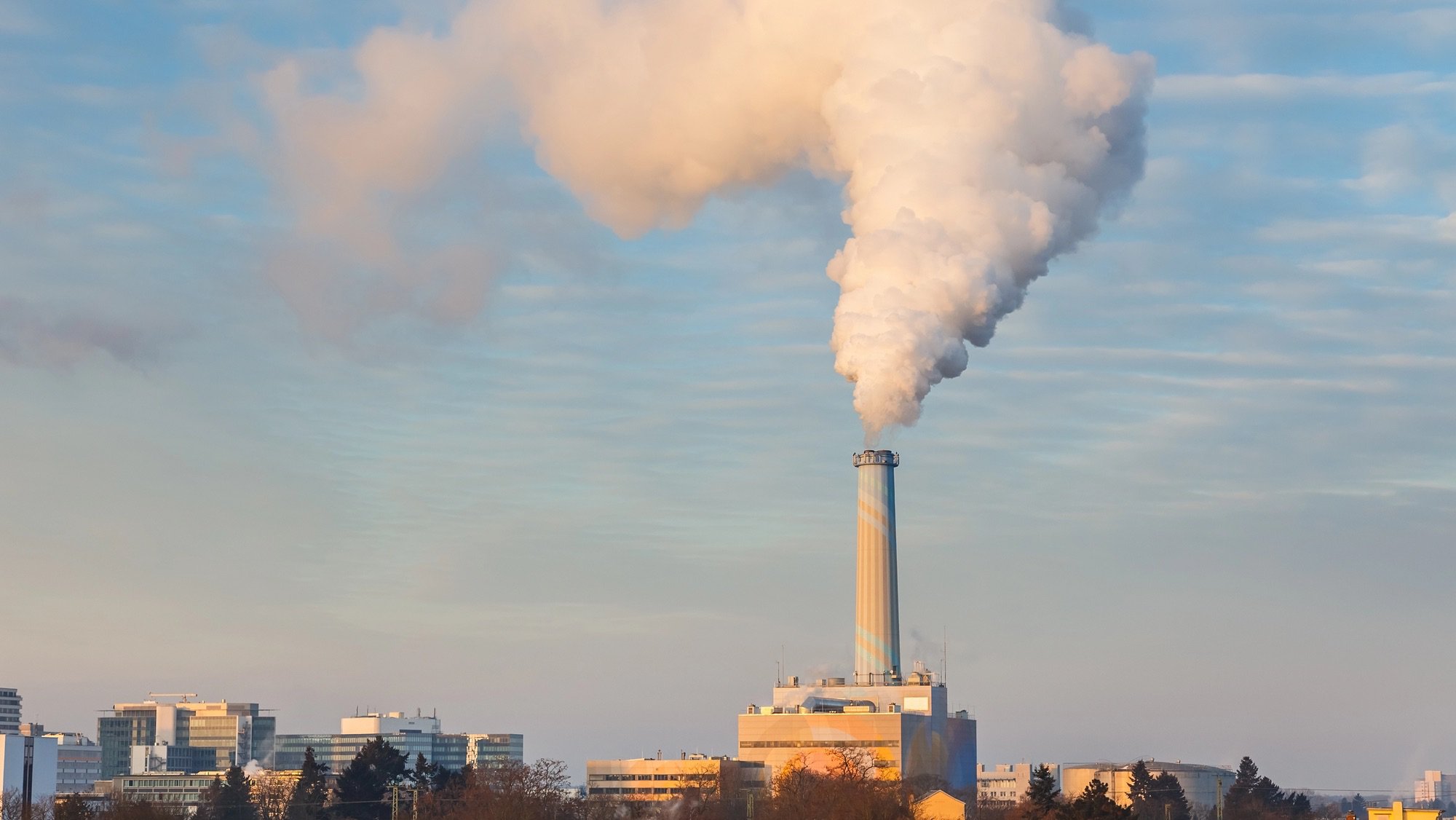 A wide shot of a tall, modern smokestack, painted in blue, white, and orange, releasing a thick, white plume of smoke into a light blue sky with wispy clouds. The smokestack is the focal point, rising above a skyline of smaller, modern buildings and trees in the foreground.
