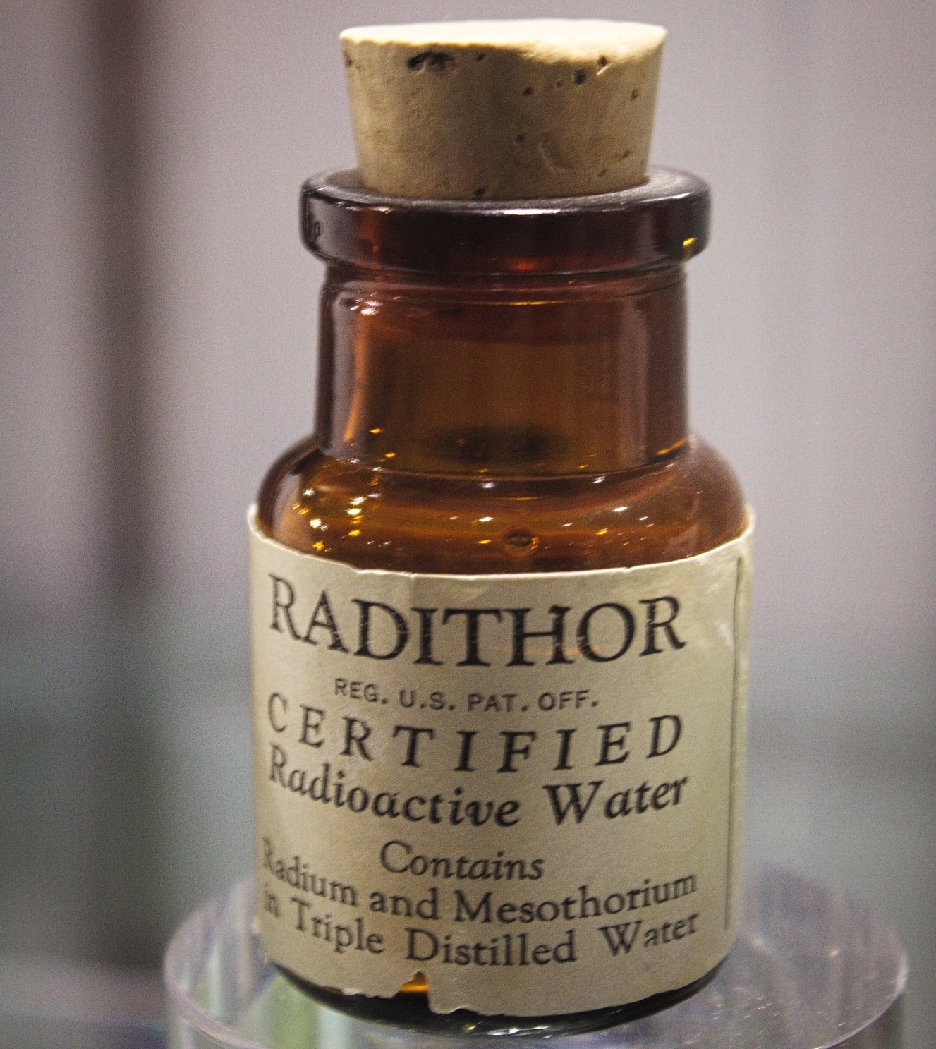 A close-up photograph of a small, amber glass bottle sealed with a cork stopper. A vintage label on the front reads "RADITHOR" in large letters, followed by "CERTIFIED Radioactive Water" and "Contains Radium and Mesothorium in Triple Distilled Water." The bottle is displayed on a clear, cylindrical pedestal.