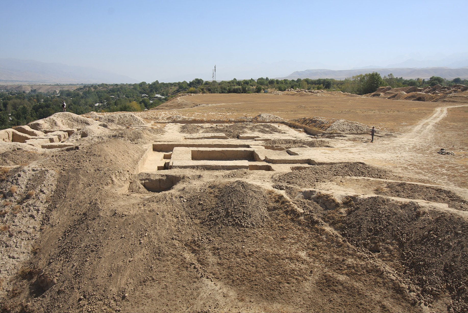 An arid archaeological site with a large, excavated rectangular foundation in the foreground, partially dug into a dirt mound. The site is surrounded by dry, barren land and hills in the distance. A few people are visible, providing a sense of scale, and the background shows a valley with green trees and a clear, hazy sky.