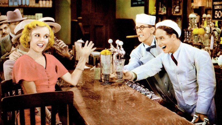 A colorized vintage photo shows a smiling blonde woman in a salmon-colored dress waving her hand while seated at a dark wooden counter or bar. Two men in white staff uniforms and hats are leaning over the counter, laughing with her, one of whom is holding a cigar. The other offers her a drink in a tall glass. Several other men in hats are visible in the background.
