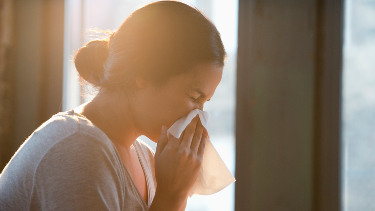 a woman sneezing by a window with sun streaming in