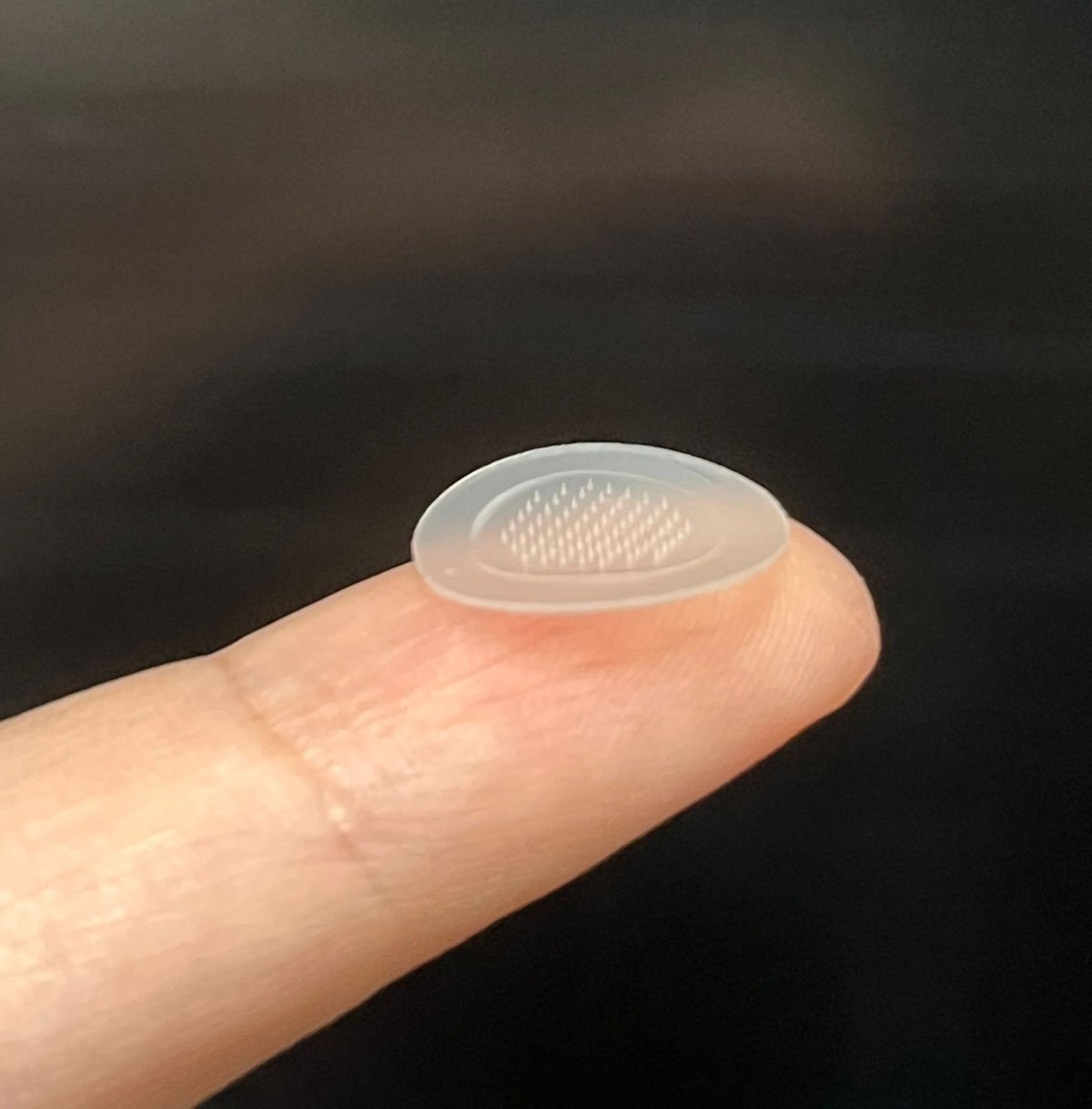 a finger holding a new pimple patch. it is circular, clear, and has tiny spikes on it