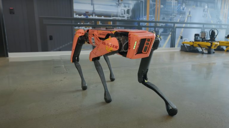 A four-legged orange robot dog from Boston Dynamics, named Spot, stands on a shiny concrete floor indoors. The robot has black legs and decals with words like “BOSTONER” and “RUSSELL” on its body. In the background, several yellow robot dogs are lined up against a wall with an industrial mural.