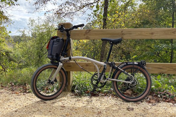 A Traildust White Brompton Electric G Line folding ebike leaning against a wooded guardrail on the C&O Towpath, green grass and trees in the background