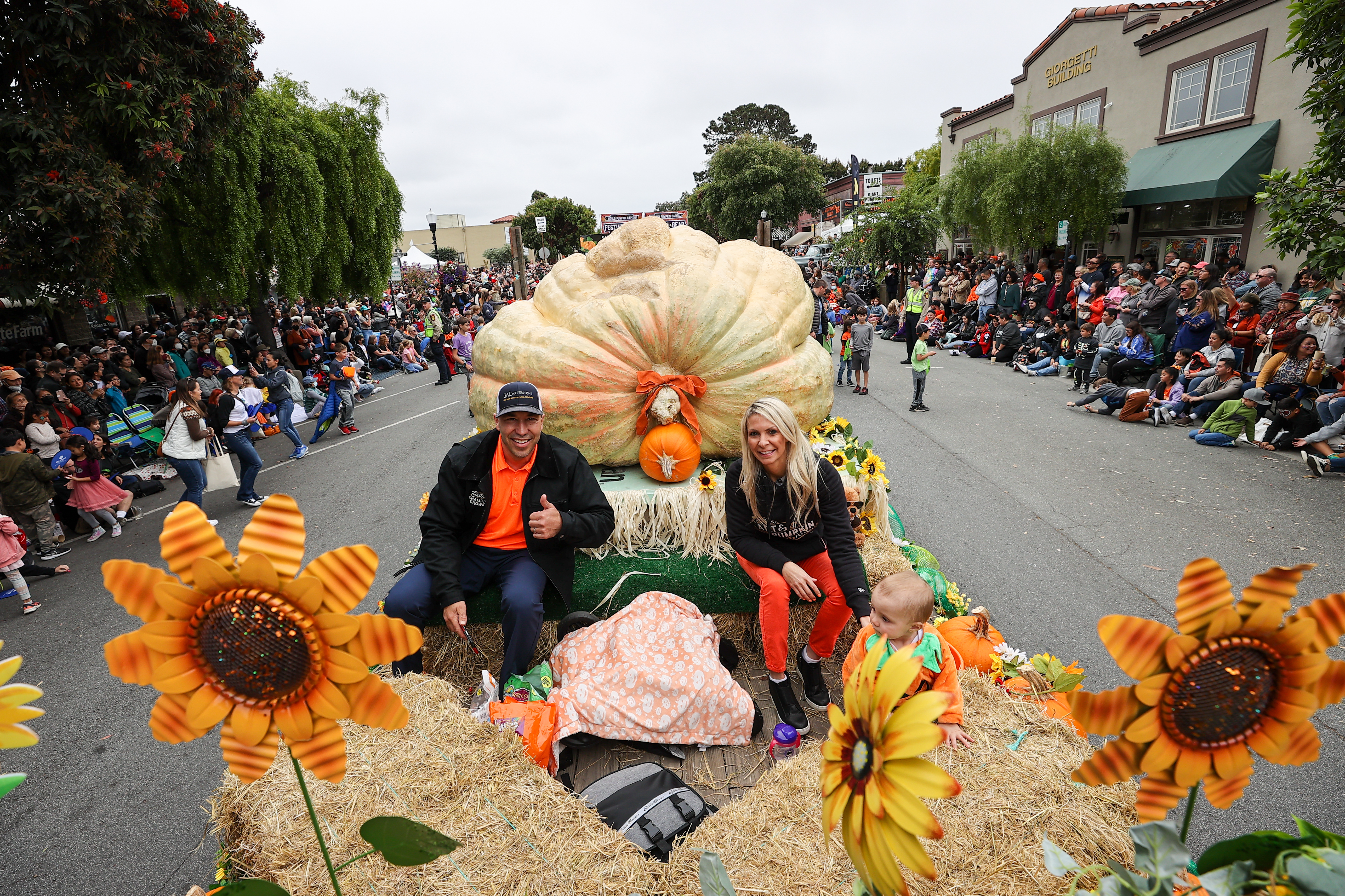 A high-angle photograph shows a giant, pale yellow pumpkin being pulled on a parade float through a street lined with a large crowd of spectators. A man, a woman, and a baby are seated on hay bales in front of the enormous pumpkin, with the man giving a thumbs-up. The float is decorated with large, orange artificial sunflowers.