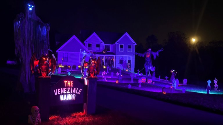 A nighttime photograph shows a suburban house transformed into a dramatic haunted house display, illuminated by intense purple and blue lighting. The front lawn is covered with numerous Halloween decorations, including glowing ghosts, demonic gargoyles flanking a sign that reads "THE VENEZIALE MANOR," skeletons, and lighted pumpkin figures. A bright moon or spotlight shines in the upper right.