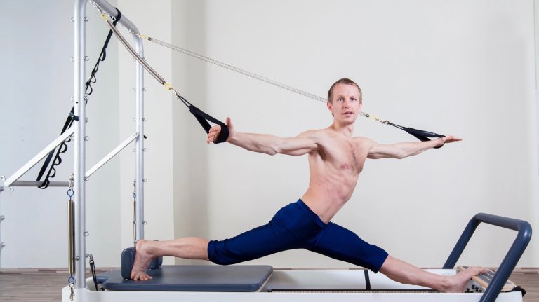 A fit, shirtless man performs a Pilates exercise on a reformer machine. He is in a deep lunge position, with his arms stretched out to the sides holding the machine's ropes. His body is taut, and he is looking straight ahead in a bright, modern studio.