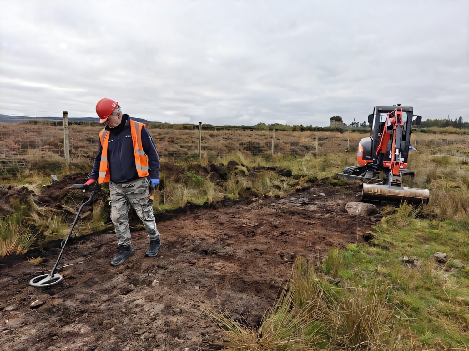 Archaeologist using metal detector in Scottish highlands with excavator tractor behind him