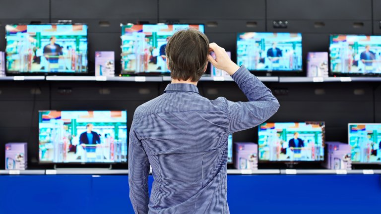 Back angle of man scratching head and looking at rows of TVs in electronics store