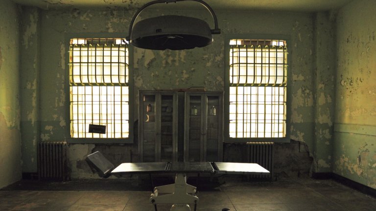 A photograph of an old, abandoned operating room with peeling, distressed green paint on the walls. The room is symmetrically framed, featuring a central metal operating table and a large surgical lamp hanging from the ceiling. Two tall, barred windows on either side are brightly backlit, casting a dramatic light into the dim room.