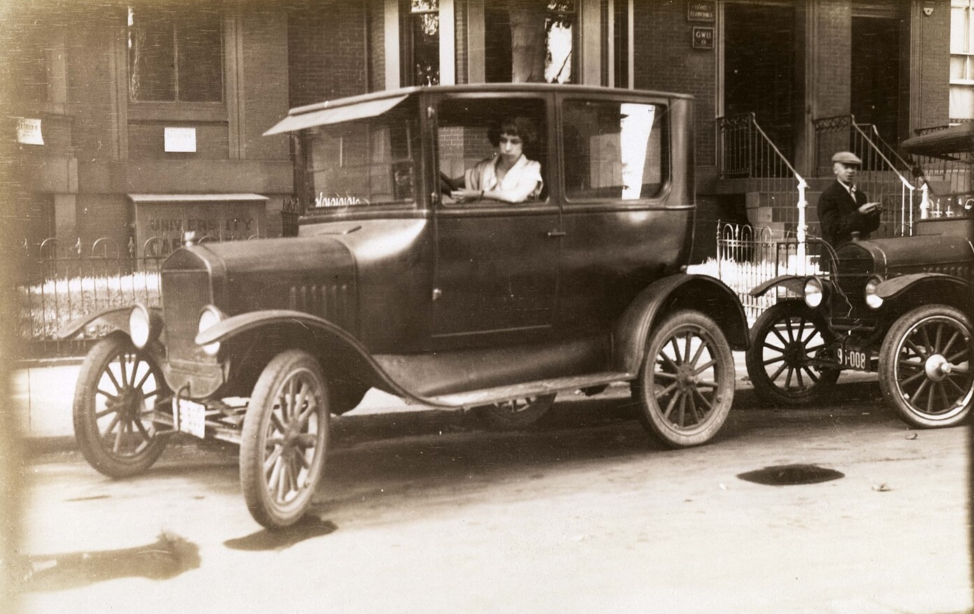 A faded, vintage photograph shows a woman, possibly Thelma Hunt, seated in the driver's seat of a black, early 20th-century automobile with the window down. She is looking forward, and her arm is resting on the door. The car is parallel parked on a city street in front of brick buildings, and a man in a cap is standing next to another vehicle on the right.