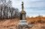 A stone monument topped with a statue of a Civil War soldier standing with his arms crossed. The monument has a dark plaque and is set upon a large rock base surrounded by dry, tall, reddish-brown grass and scrub. Bare trees stand in the background under a cloudy sky.