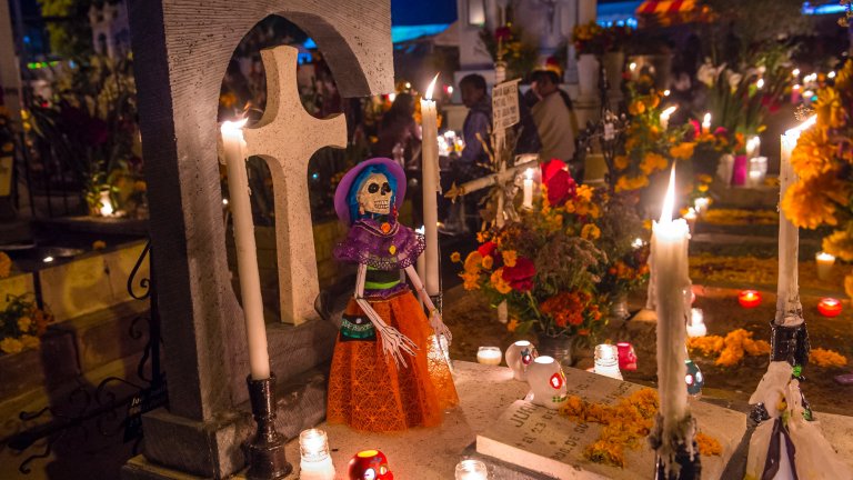 A dark, vibrant nighttime scene in a cemetery during a Day of the Dead (Día de Muertos) celebration. In the foreground, a decorative, brightly colored skeleton doll (Catrina) stands next to a stone grave marker topped with a cross. The grave is covered in bright orange marigold flowers, and numerous lit white candles and small tea lights illuminate the scene, casting a warm glow on flowers and blurred figures in the background.