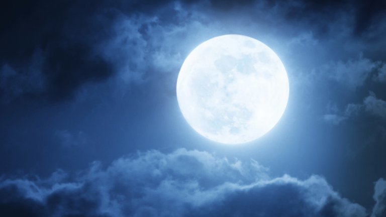 A striking photograph of a bright, full moon against a dark, deep blue night sky, partially obscured by dark, dramatic clouds. The moon is intensely bright, giving the scene an ethereal glow.