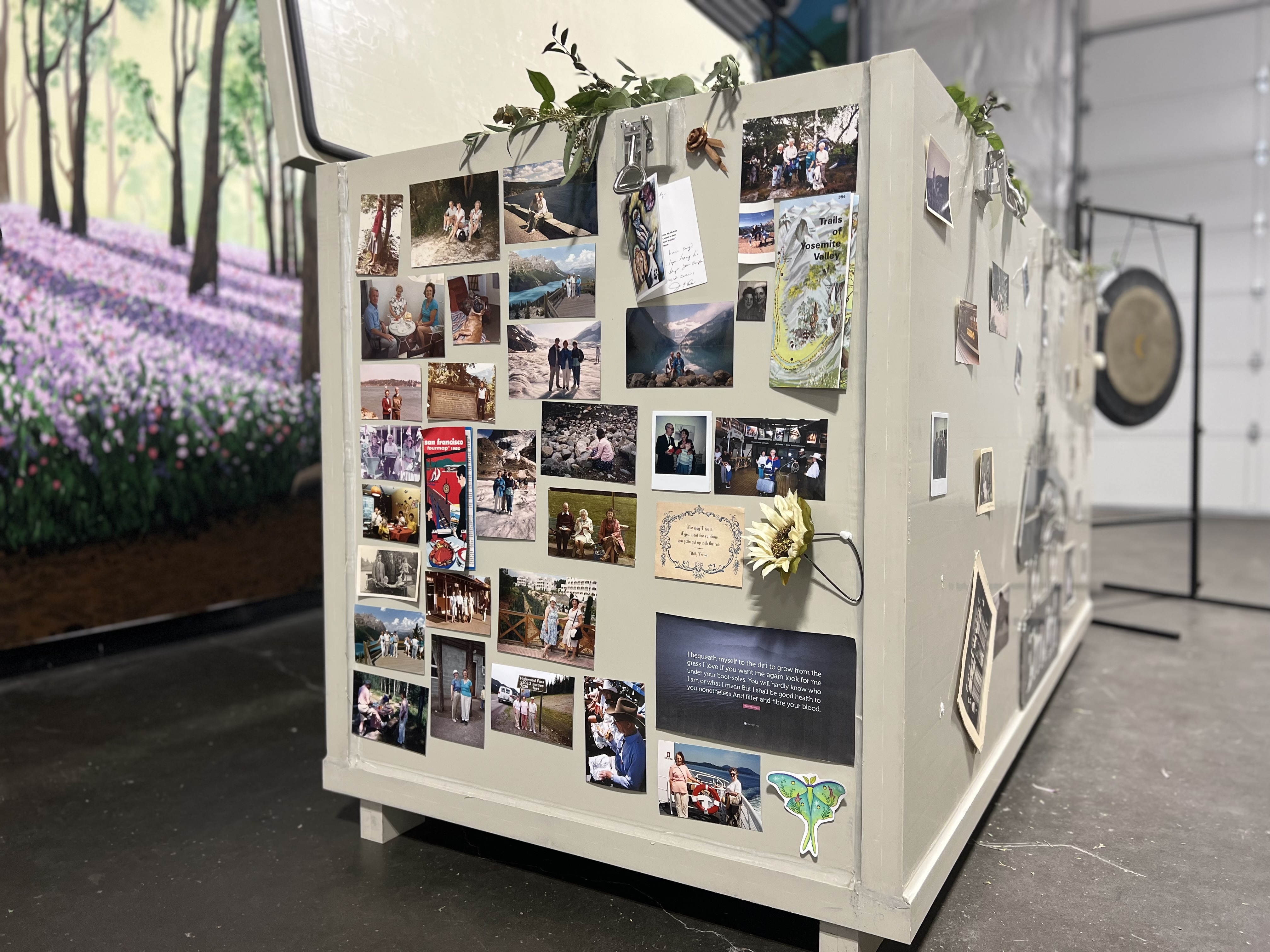 A photograph of a large, pale-gray, rectangular composting vessel. The side of the vessel is decorated with dozens of personal photos, postcards, and small mementos, attached with magnets. In the background on the left is a colorful mural depicting a forest with purple flowers, and a large metal gong is visible on a stand on the right. The floor is dark concrete.