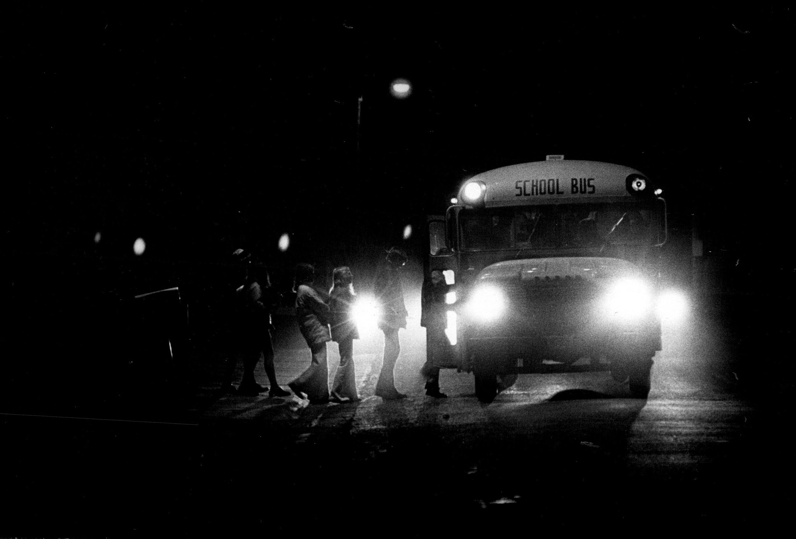 A grainy, black and white photograph captured at night shows the headlights of a school bus and a car illuminating a small group of indistinct figures, likely children, standing near the bus door. The sign "SCHOOL BUS" is visible above the windshield, and the overall scene is very dark, suggesting a very early hour.