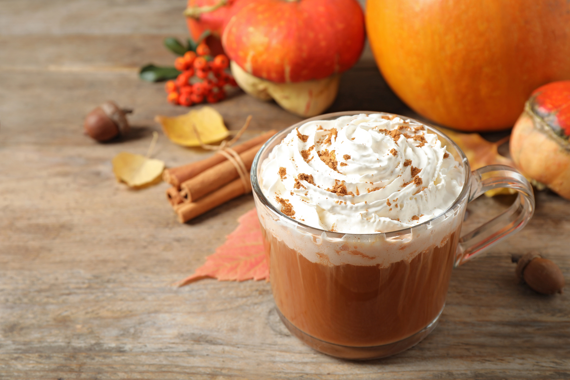 A warm, autumnal still life featuring a clear glass mug of a pumpkin spice latte topped with whipped cream and sprinkled with cinnamon. It sits on a rustic wooden surface surrounded by fall decorations, including cinnamon sticks tied together, fallen leaves, acorns, rowan berries, and small pumpkins.