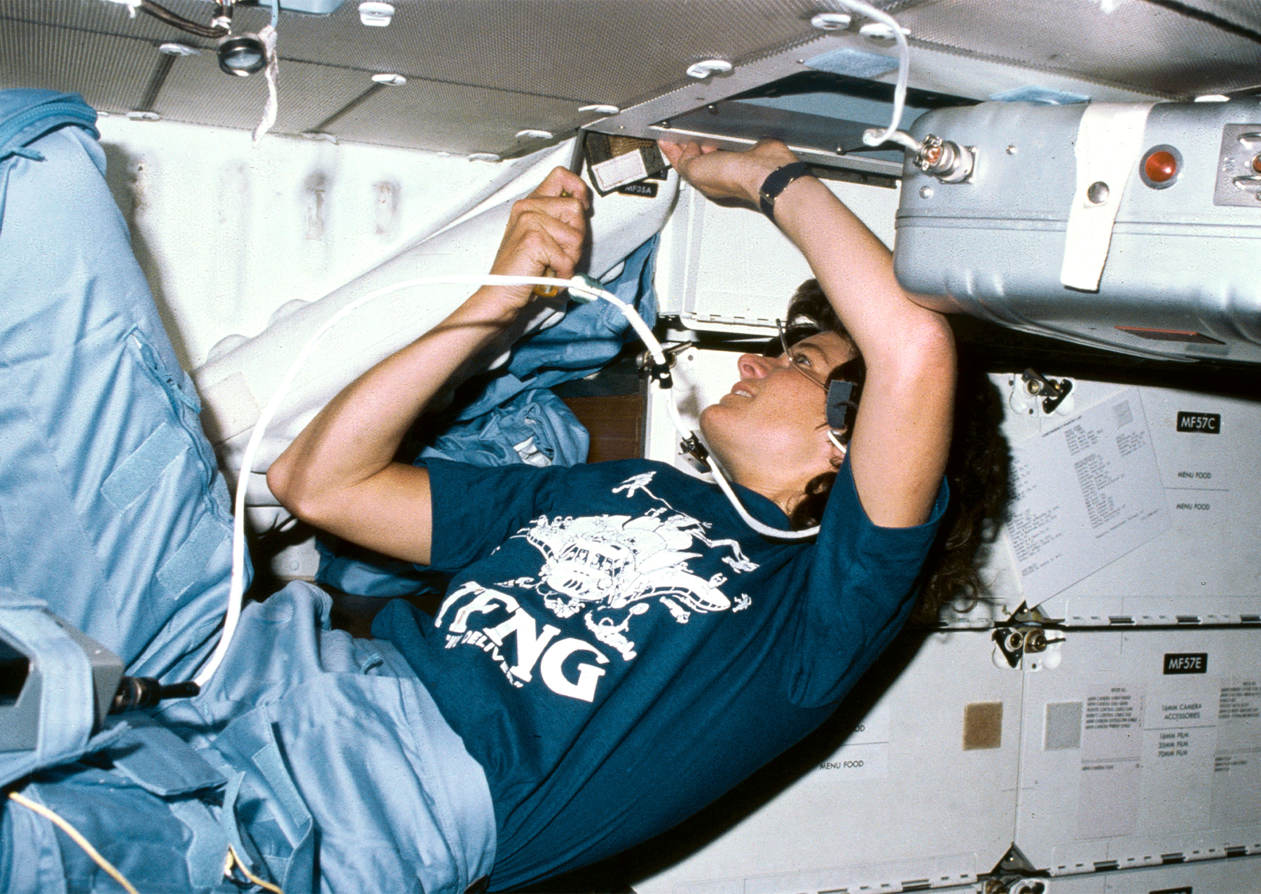 Sally Ride wearing a "TFNG" shirt as she cleans an air filter system during her 1983 flight aboard Space Shuttle Challenger. TFNG stands for Thirty-Five New Guys, the nickname that the 1978 class of astronaut candidates gave themselves.