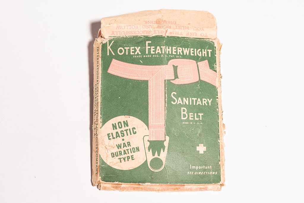 A faded, worn, vintage cardboard box for a Kotex Featherweight Sanitary Belt. The box has a green background with tan and black text and an illustration of a pink T-shaped belt. A circular inset bubble reads "NON ELASTIC WAR DURATION TYPE." The box is slightly open at the top, and the edges are damaged.