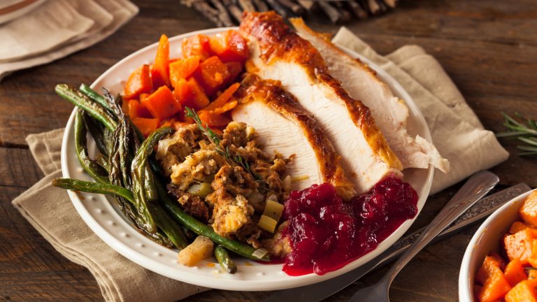 A plate filled with a traditional holiday meal, featuring sliced roasted turkey breast, a scoop of stuffing, green beans, diced sweet potatoes, and a spoonful of cranberry sauce, all resting on a white plate set on a rustic wooden table.