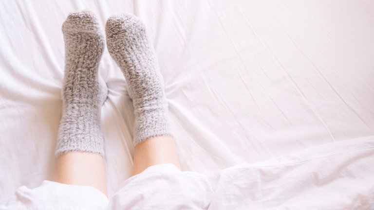 A person's lower legs and feet, wearing thick, gray fuzzy socks, are stretched out against a clean white duvet or sheet. The image is bright and minimal, suggesting a cozy, relaxed moment in bed.
