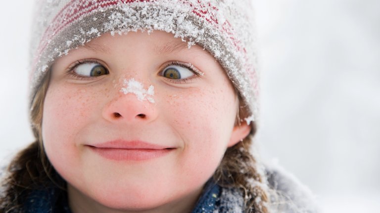 A close-up photograph of a young child's face with freckles, wearing a knit winter hat dusted with snow. The child is smiling and deliberately crossing their eyes. The background is a blurry white winter scene.