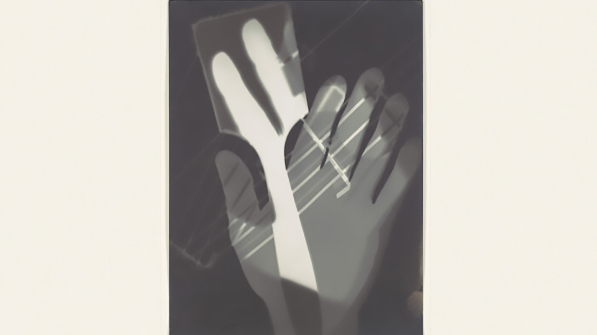 A black and white photograph, likely a photogram, shows a stylized image of a human hand overlayed with a partially opaque rectangular object. The outlines of the hand and the object are defined by areas of white and gray, creating a dramatic, X-ray-like effect against a dark background.