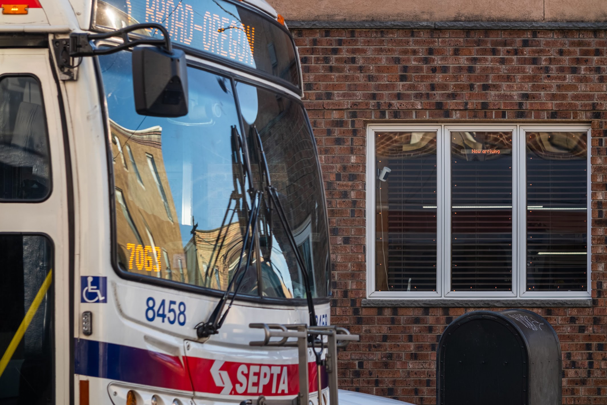 A close-up photograph of the front of a white SEPTA public transit bus parked on a city street. The bus's windshield reflects the surrounding buildings, and a digital sign is visible near the top. The bus is next to a brick building with a large white-framed window.