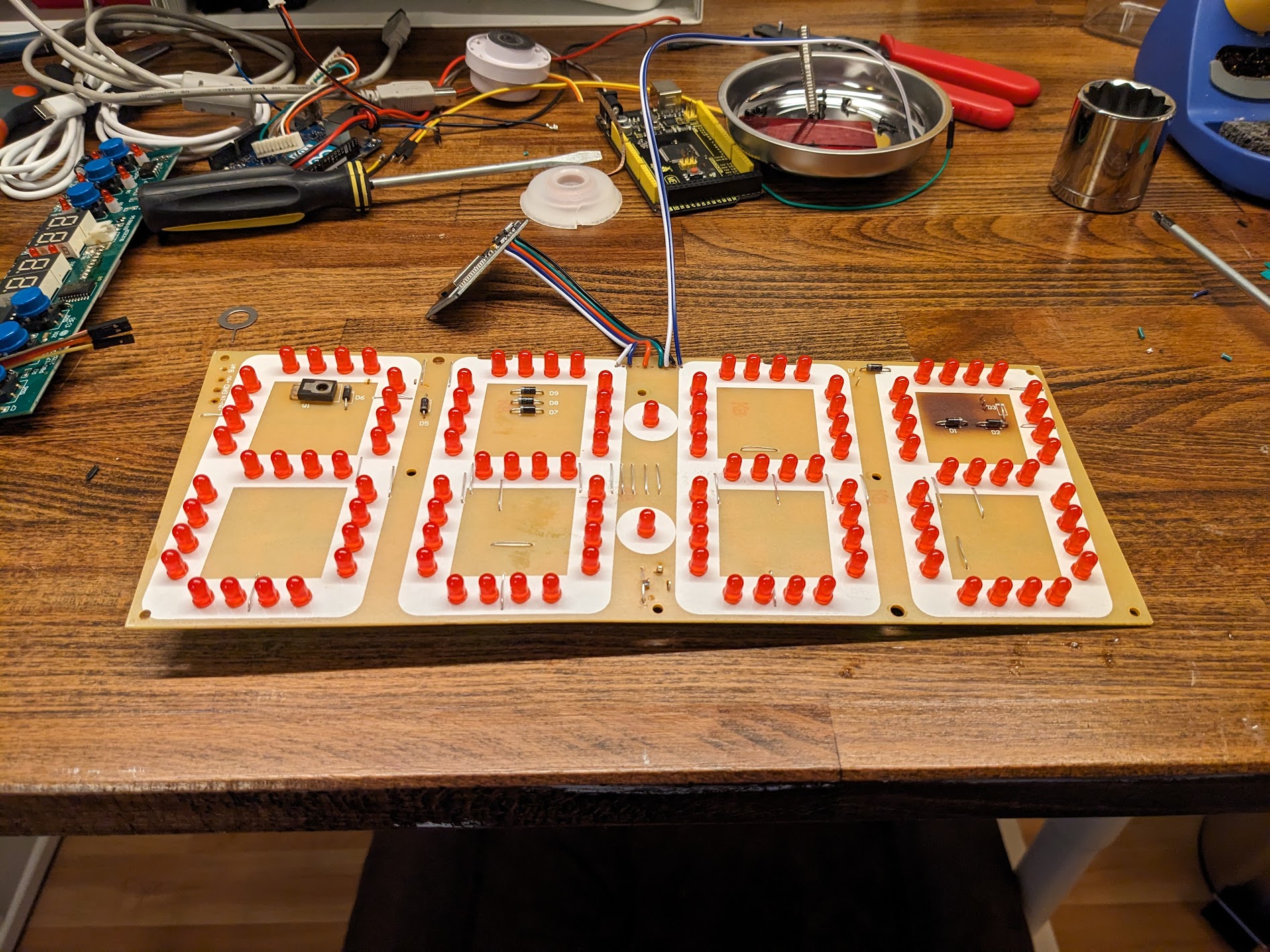 An overhead shot of an electronic circuit board laid on a wooden workbench, with dozens of small, red LED lights arranged in a large seven-segment display pattern, ready to form numbers or letters. The surrounding area is cluttered with wires, tools, a soldering station, and other electronic components.