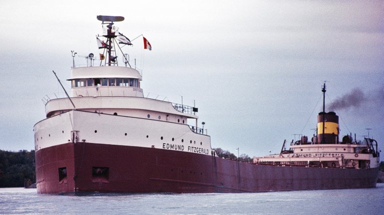 A historical color photograph of the large Great Lakes freighter, the SS Edmund Fitzgerald, sailing on the water. The ship has a prominent white superstructure at the bow with a dark red hull and a tan smokestack near the stern emitting a plume of black smoke. The weather appears overcast.