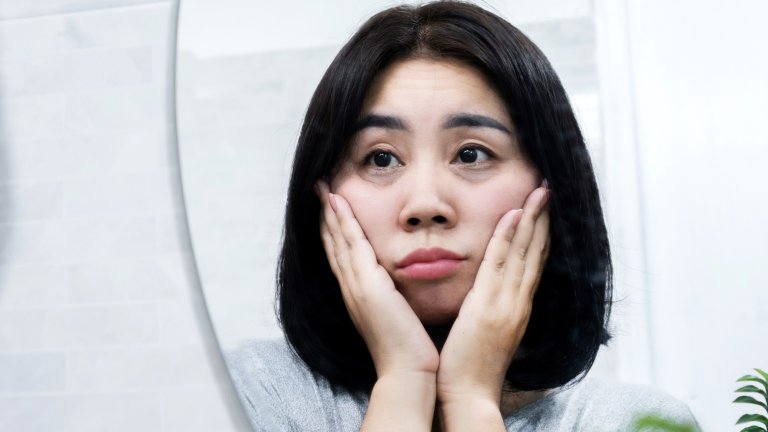 A close-up shot of an Asian woman with dark hair looking intently into a mirror, touching both sides of her face with her hands. Her expression is concerned or thoughtful, and she appears to be examining the skin or puffiness under her eyes. The background is bright and dominated by the reflection of a bathroom or vanity.