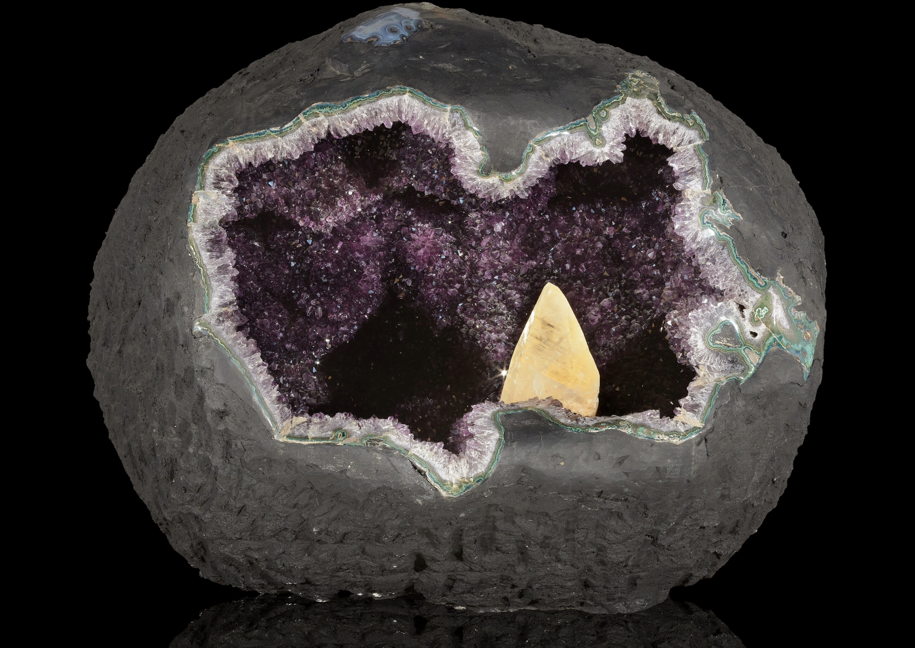 This amethyst geode with calcite crystal from Uruguay is truly a giant gem. It weighs in at 1,933 pounds and is 44 inches long. The calcite crystal stands 10 inches high and the amethyst crystals are considered gem quality.  Geodes like this one originate within volcanic vugs, or hollow cavities that were created by ancient gas bubbles within solidified lava. Over thousands of years, mineral-rich hydrothermal fluids percolated into these voids, bringing in layers of iron and silica that crystallized under the immense heat and pressure from Earth’s core and mantle to form the amethyst’s purple hue. I