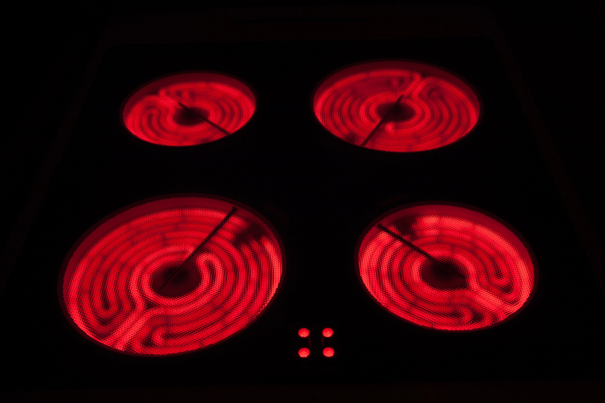A dark, overhead photograph of a glass-ceramic radiant electric stovetop in use. Four circular cooking zones are glowing intensely red with visible heating elements, contrasted against the black surface in a dark setting.