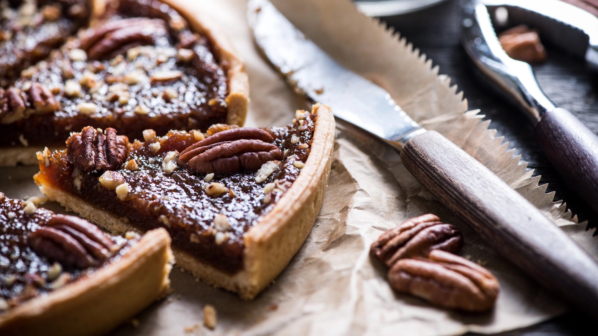 A close-up shot of several slices of pecan pie with a rich, dark filling and whole pecans arranged on top. The pie slices are resting on wrinkled brown paper, next to a silver knife with a serrated edge and a nutcracker. Two shelled pecans are visible in the foreground.