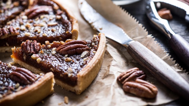 A close-up shot of several slices of pecan pie with a rich, dark filling and whole pecans arranged on top. The pie slices are resting on wrinkled brown paper, next to a silver knife with a serrated edge and a nutcracker. Two shelled pecans are visible in the foreground.