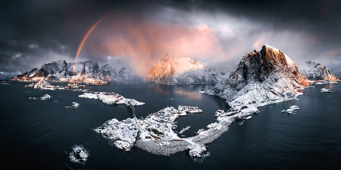 the sun rises over snow capped white mountains. a rainbow is surrounding one of the mountains