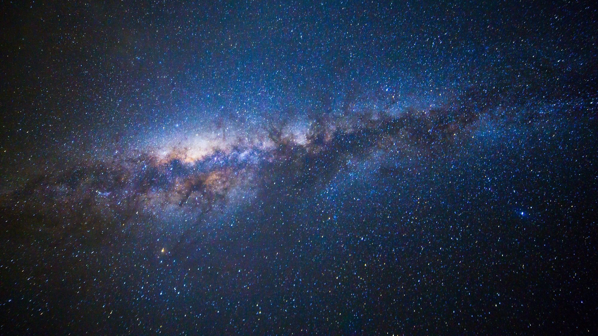 A wide-field astronomical photograph of the night sky showing the bright, luminous band of the Milky Way Galaxy stretching diagonally across a dark expanse filled with countless stars.