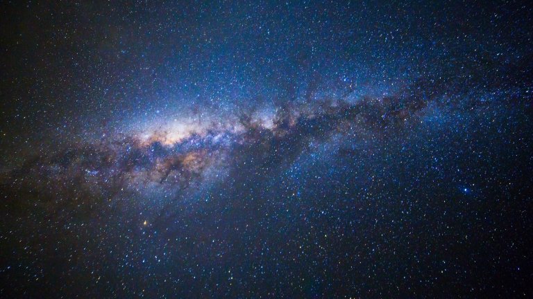 A wide-field astronomical photograph of the night sky showing the bright, luminous band of the Milky Way Galaxy stretching diagonally across a dark expanse filled with countless stars.