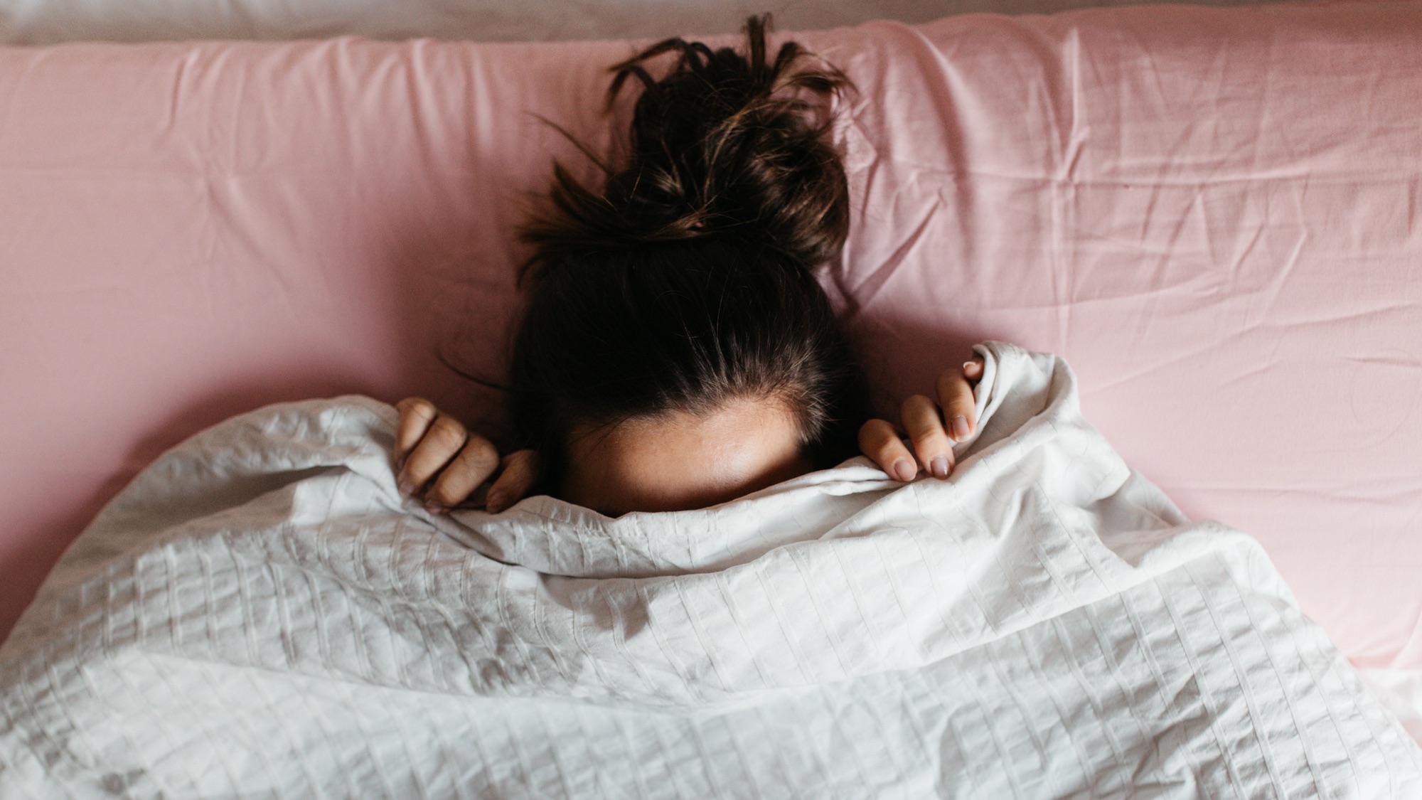 Playful young woman hiding face under blanket while lying in cozy bed, pretty curious girl feeling shy peeking from duvet, covering with white sheet, head shot close up. Top view