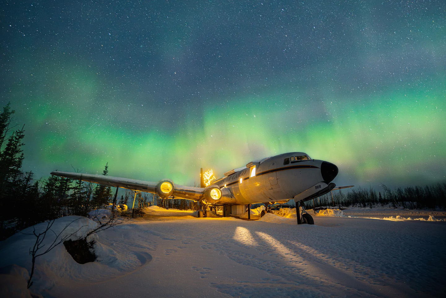 An old DC-6 cargo plane converted into lodging rests in the snow under a glowing aurora, its windows and engines illuminated against a dark, starry winter sky.