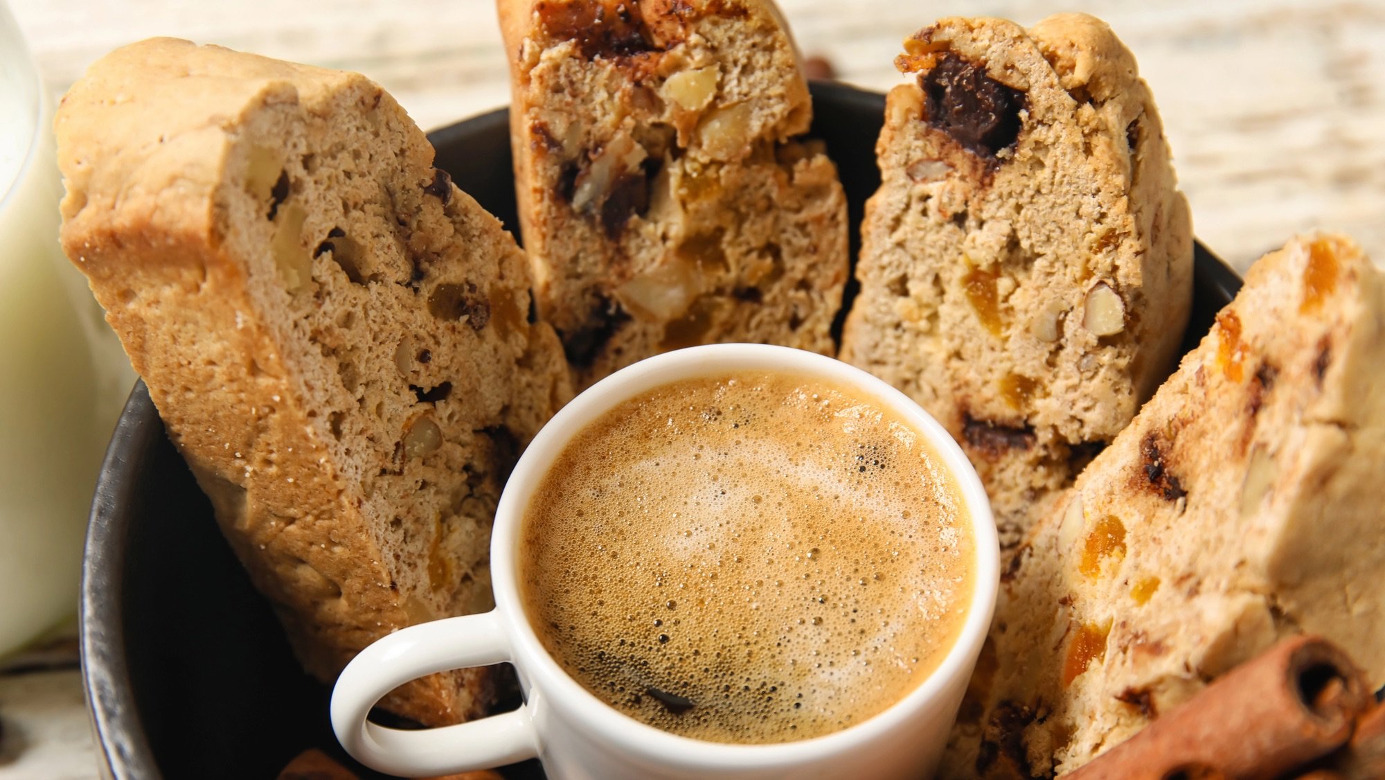 A close-up, high-angle view of four biscotti pieces standing upright in a dark bowl, surrounding a small white cup filled with espresso topped with foam. The biscotti are thick, light brown, and contain visible chunks of nuts, dried fruit (possibly apricot or orange peel), and dark chocolate. A cinnamon stick is visible in the bottom right corner, and a glass of milk or cream is partially visible on the left. The background is a blurred light wooden surface.