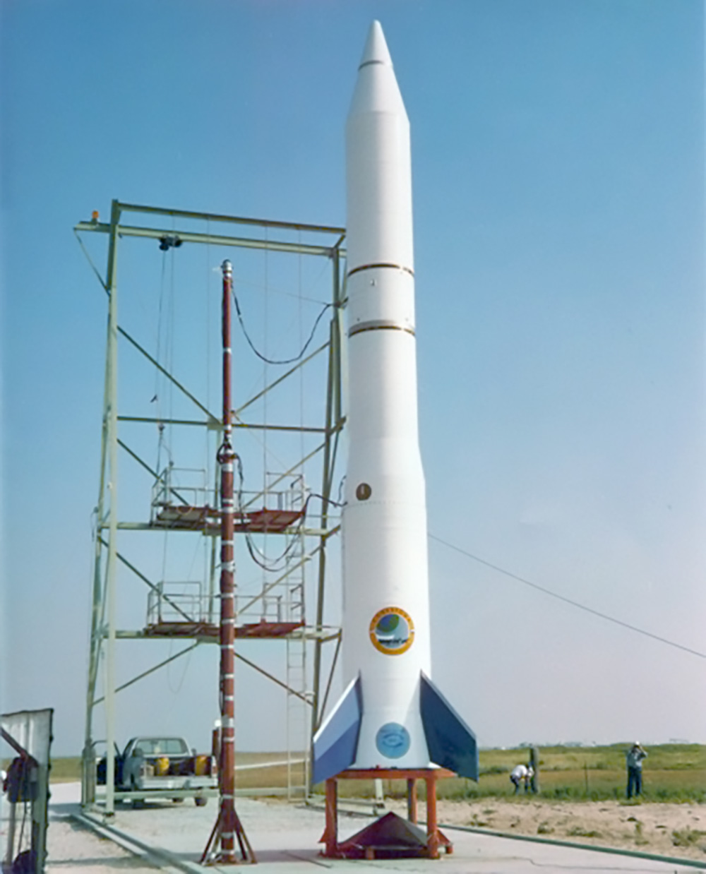 A tall, slender, white rocket with blue tail fins, featuring a circular blue and gold insignia on its body, stands upright on a launch pad. A metal service structure or tower is positioned next to it. In the background, there is a flat, grassy field and a clear blue sky. A pickup truck is visible near the base of the tower, and a few individuals are standing in the distance to the right of the rocket. The image title suggests this is the Conestoga I rocket prepared for launch.