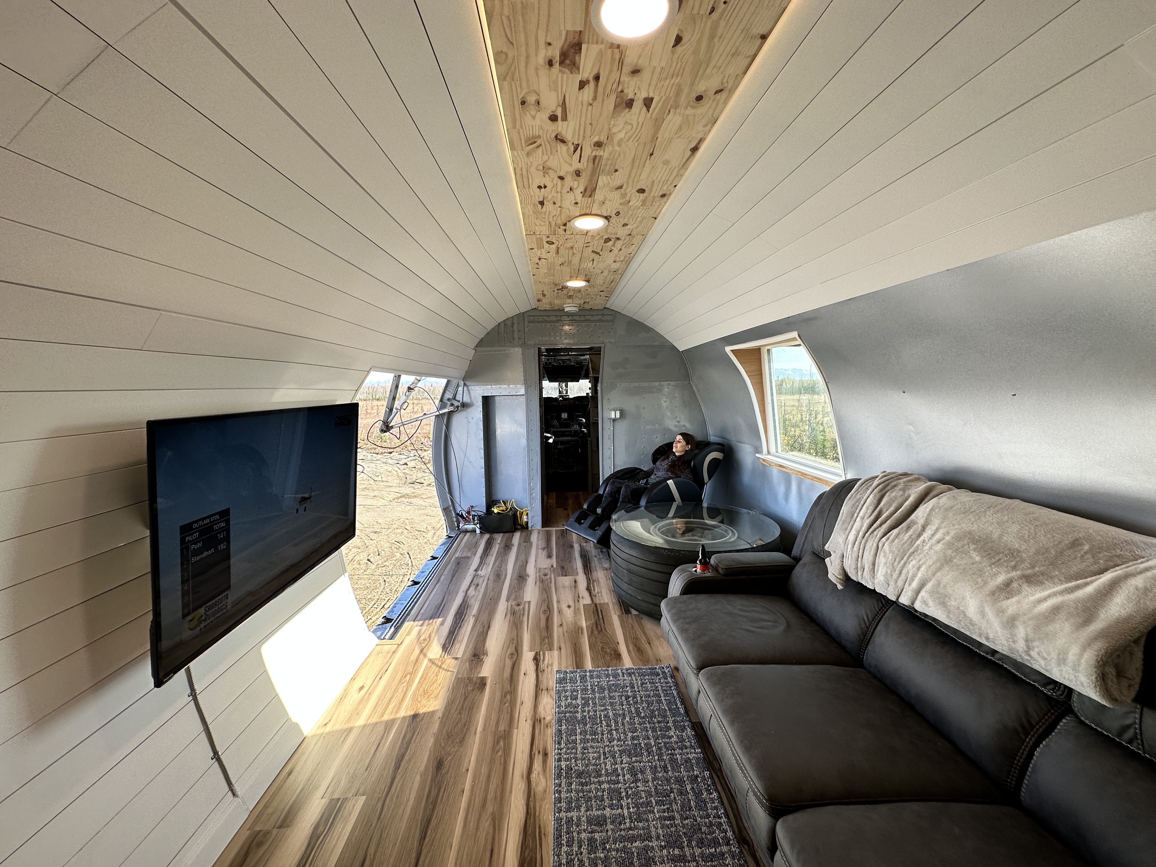 A wide-angle photo showing the completed interior of a renovated aircraft fuselage, now serving as a modern living space. The walls and arched ceiling are lined with white shiplap paneling, accented by a central wooden strip. The room features hardwood-style flooring, a large dark couch, a flat-screen TV mounted on the wall, and a person relaxing on the couch.