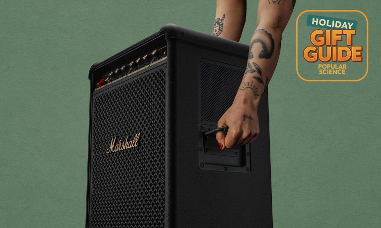 Two tattooed arms lifting a black Marshall Bromley 750 party speaker up by its handles against a green holiday gift guide background