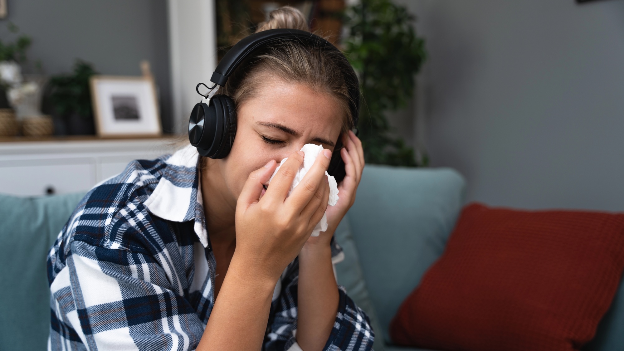 Young sad girl sitting alone at home on sofa listening to sad music on wireless headphones crying and wiping her nose with a paper tissue.