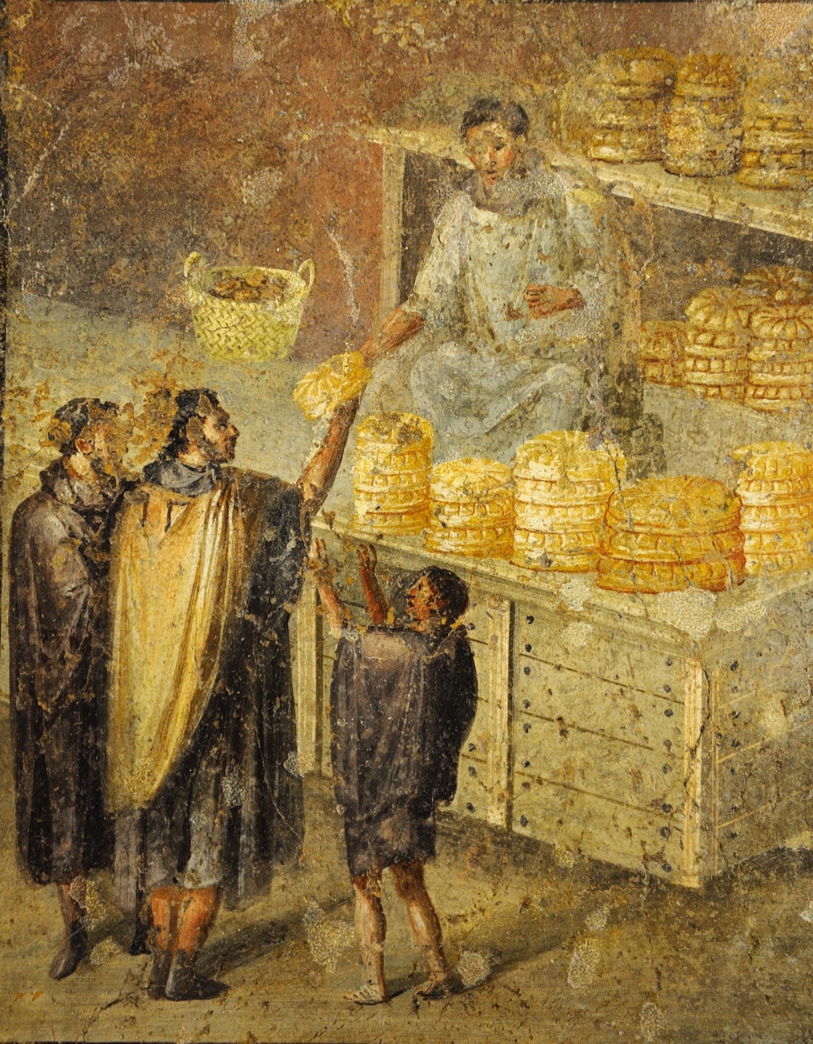An ancient Roman fresco depicts a scene from a bakery or bread stall. A vendor, seated or standing behind a counter laden with stacked, round loaves of bread, hands a piece of bread to a customer wearing dark robes. Two other figures stand to the left, one of whom appears to be a child reaching up, possibly also for bread. The bread is a distinctive, round, scored loaf typical of those found in archaeological digs in Pompeii. A small basket is visible on a surface to the left.