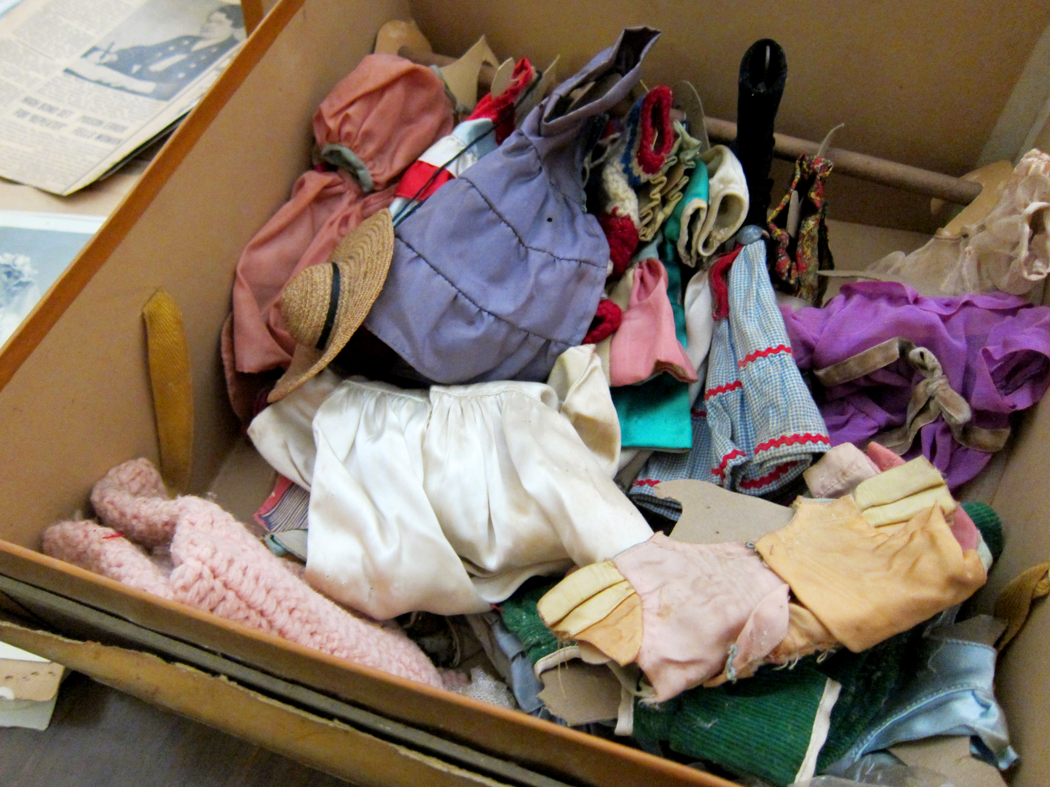 A high-angle, close-up shot of a cardboard box filled with a variety of miniature handmade outfits and accessories for Tommy Tucker the squirrel. The colorful pile includes small dresses in purple, pink, and blue, a tiny straw hat with a black band, white silk-like garments, and knitted items. In the background, a vintage newspaper clipping is partially visible.