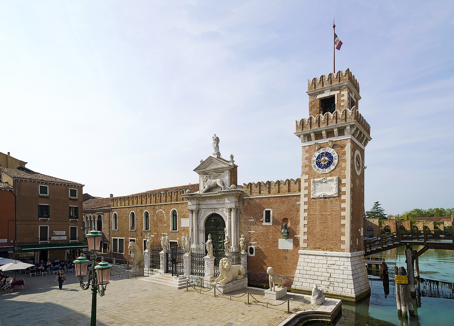 A sunny daytime view of the main land entrance to the Venetian Arsenal, known as the Porta Magna, located in the Campo de l'Arsenal. The entrance consists of a rusticated brick wall and a central marble gate topped with a statue of the Winged Lion of Saint Mark. To the right is a prominent brick tower (Torre di Porta Nuova or Porta Vecchia) with a large clock face and a crenellated top flying a flag. Two marble lions flank the gate, and additional statues are visible in the foreground near the water's edge. To the left is a square (campo) with lamp posts and an outdoor cafe area, while to the right, a wooden bridge crosses a canal.