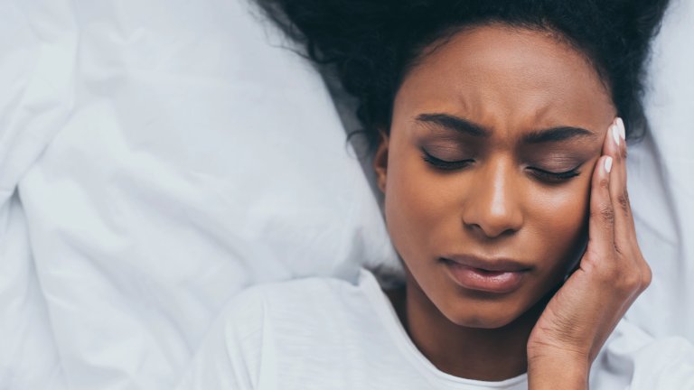 An overhead shot of a Black woman with dark curly hair lying on white bedding with her eyes tightly shut in a pained expression. She has one hand pressed against her temple, illustrating a headache or migraine.