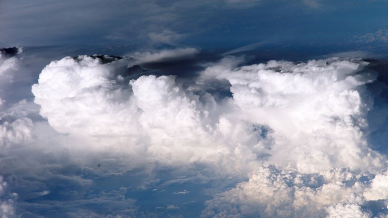 An aerial, high-altitude view looking down at a vast formation of billowing white cumulus clouds. The clouds appear thick and textured, with some peaks rising higher than others, casting soft shadows on the lower cloud layers and the dark blue atmosphere below. The composition captures the "clumping" effect of moisture in the upper atmosphere.